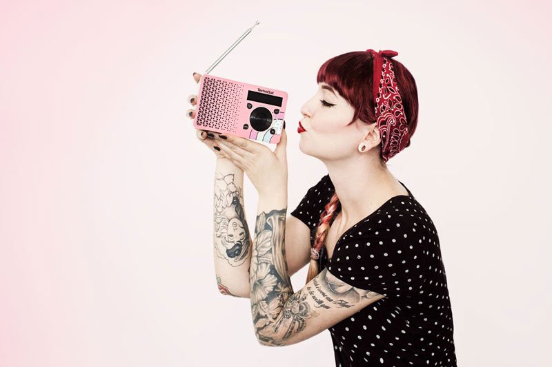 Frau mit Tattoos und rotem Bandana küsst ein pinkes Retro-Radio vor einem rosa Hintergrund. (Werbefotograf Berlin)